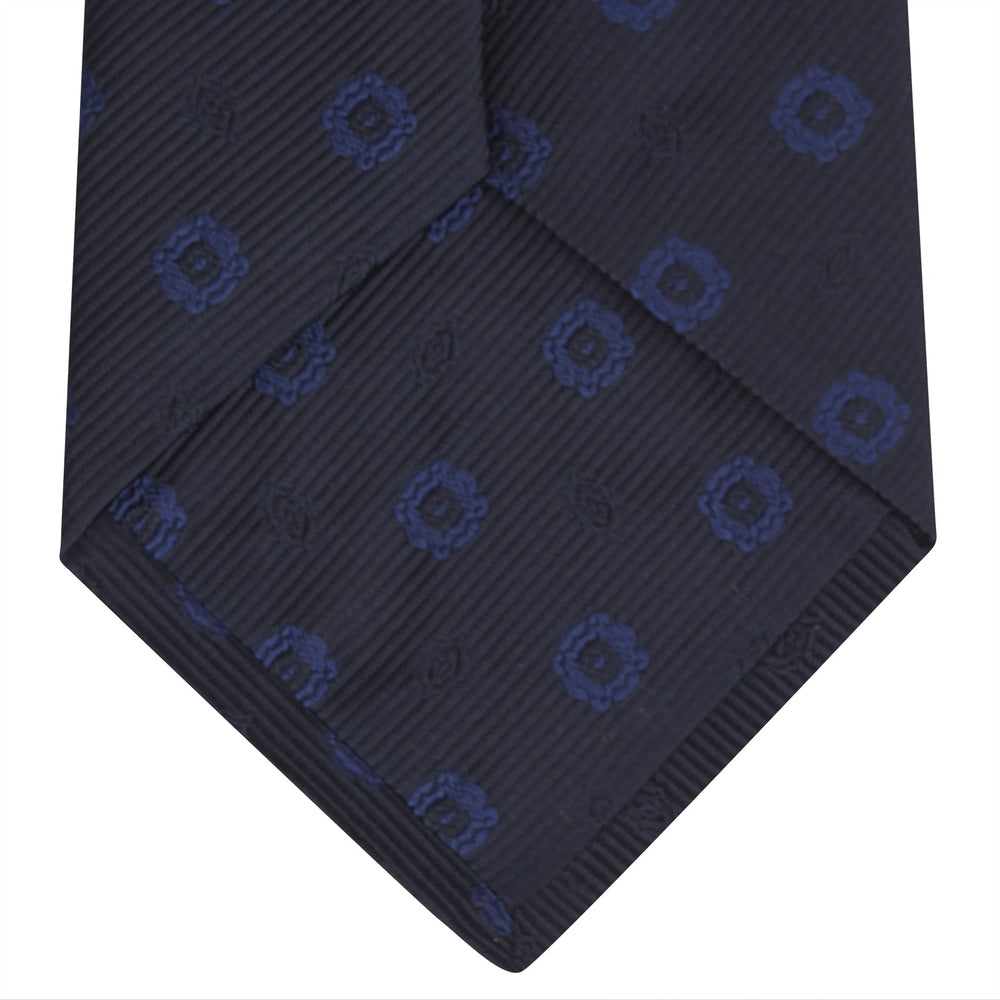 The Turnbull & Asser Navy and Blue Emblem Spot Silk Tie features a classic pattern of light blue motifs on navy silk, showcased rolled on a white background—a refined accessory for any office shirt.