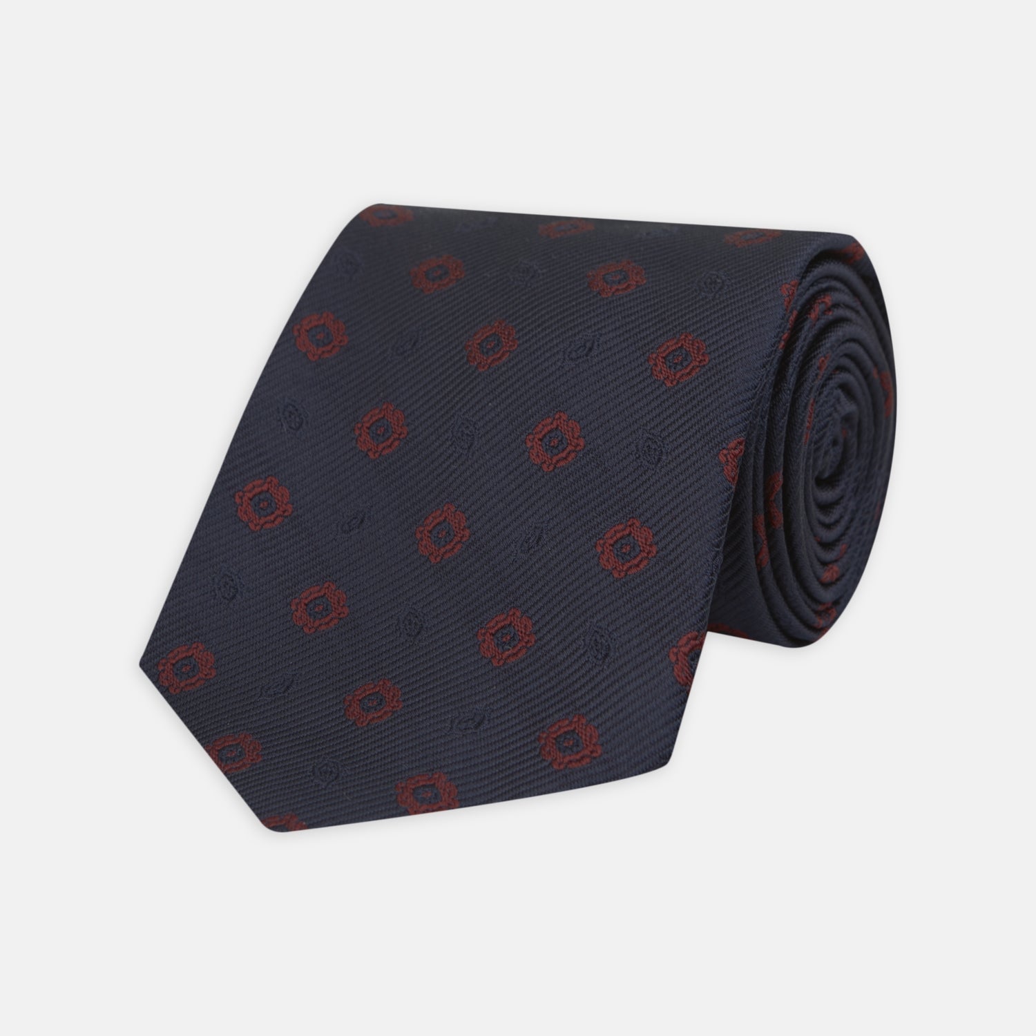 View 1 of product The Turnbull & Asser Navy and Burgundy Emblem Spot Silk Tie features a subtle textured pattern with small burgundy emblems on navy silk. Perfectly rolled with a pointed tip, it pairs well with any office shirt.