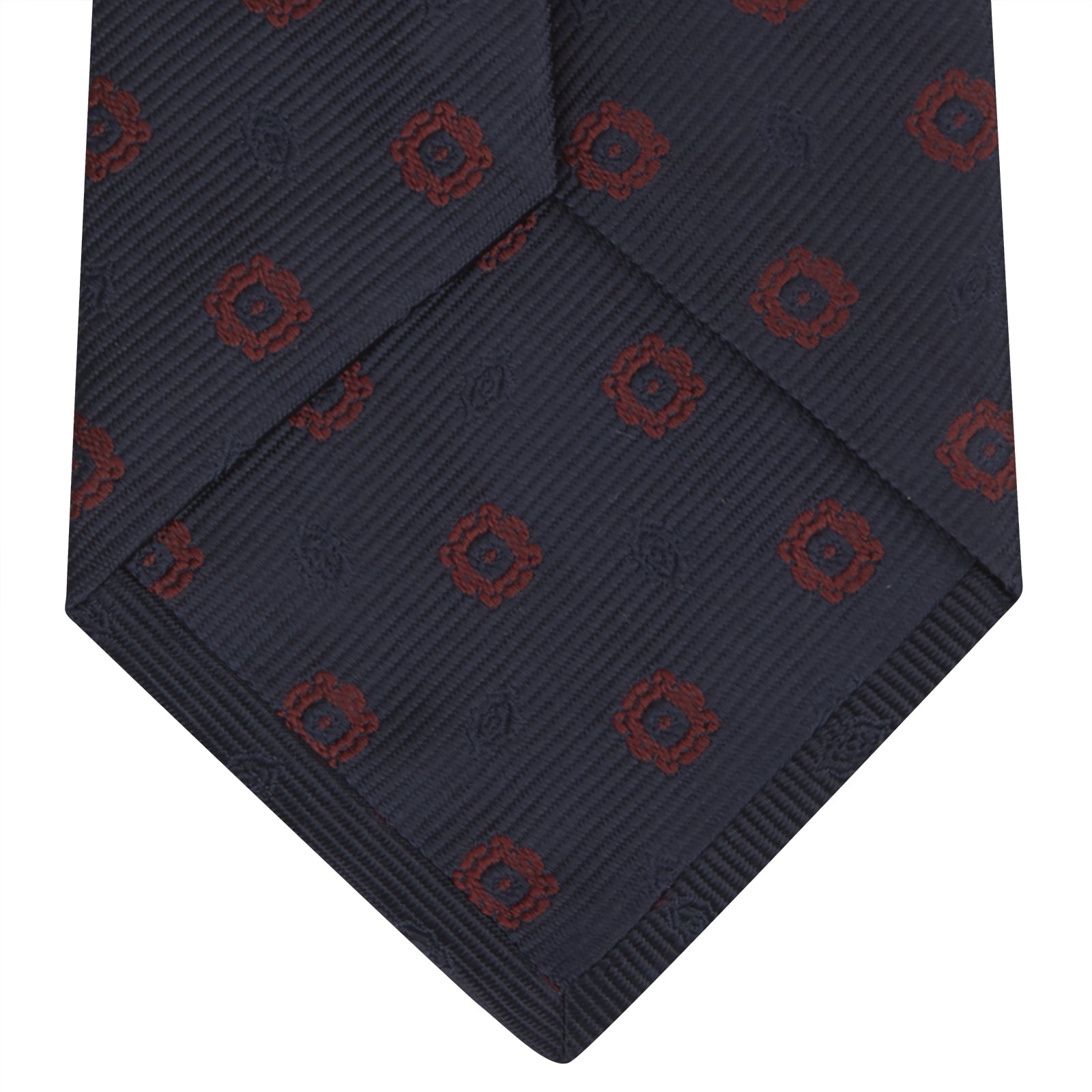 View 4 of product The Turnbull & Asser Navy and Burgundy Emblem Spot Silk Tie features a subtle textured pattern with small burgundy emblems on navy silk. Perfectly rolled with a pointed tip, it pairs well with any office shirt.