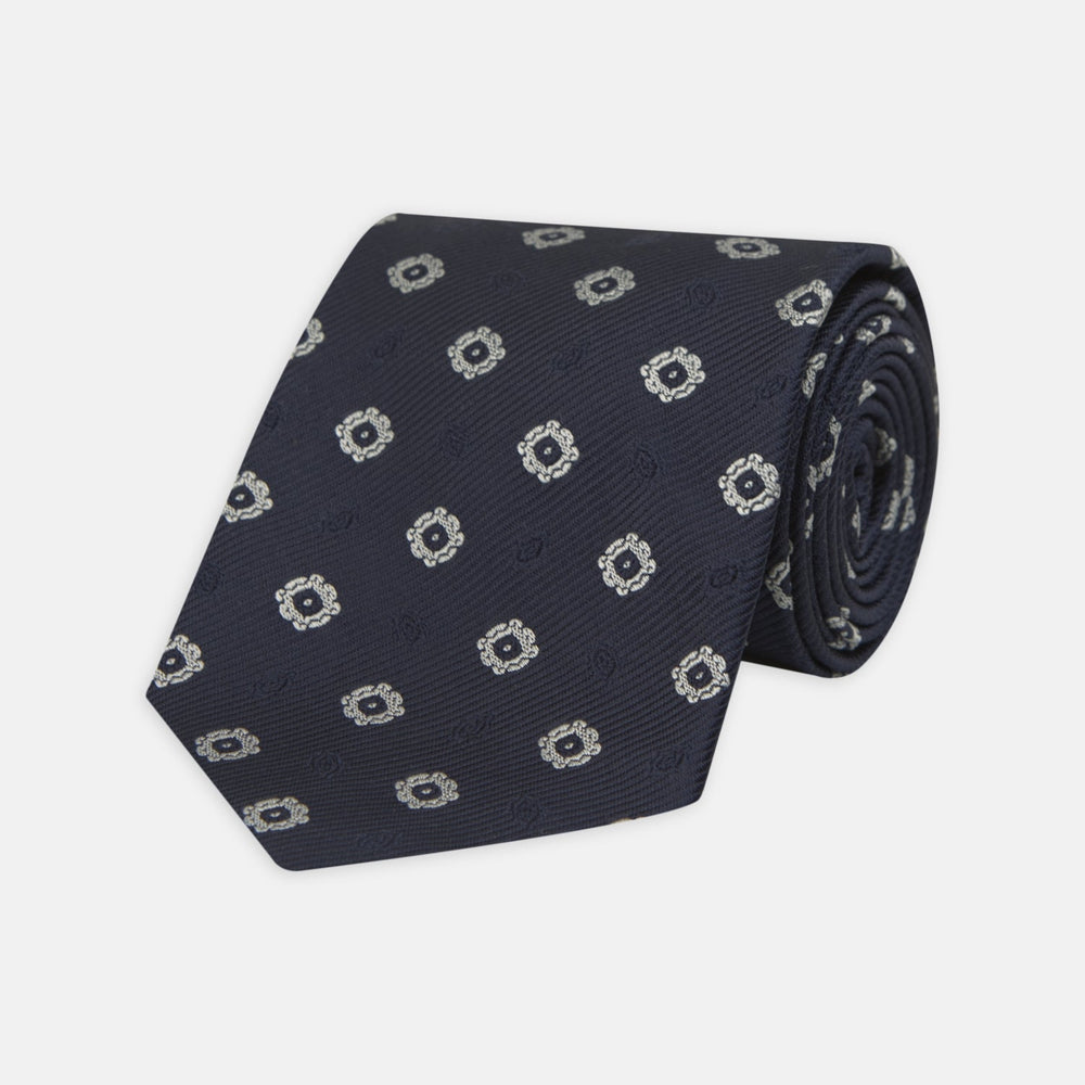 The Turnbull & Asser Navy and Silver Emblem Spot Silk Tie features a refined repeating emblem pattern in silver on navy silk, making it an elegant accessory for any office shirt.