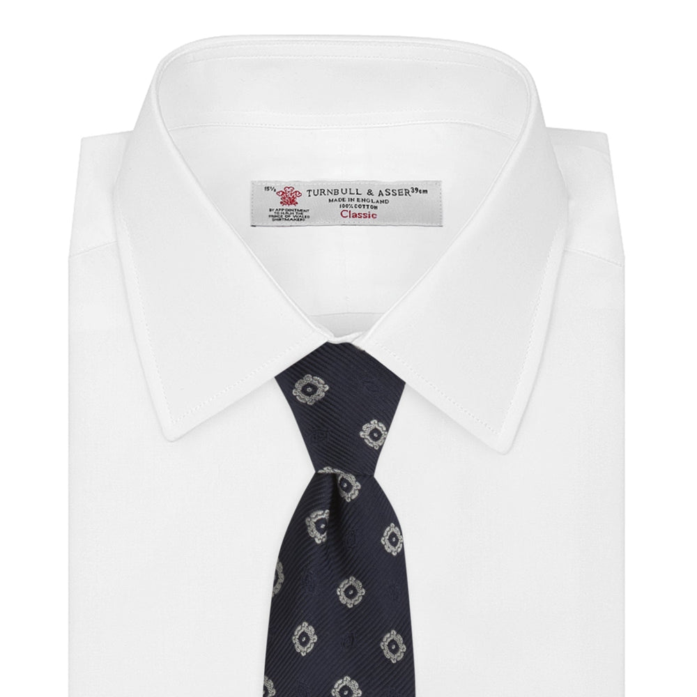 The Turnbull & Asser Navy and Silver Emblem Spot Silk Tie features a refined repeating emblem pattern in silver on navy silk, making it an elegant accessory for any office shirt.