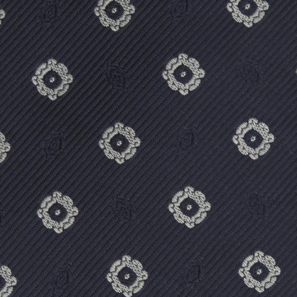 The Turnbull & Asser Navy and Silver Emblem Spot Silk Tie features a refined repeating emblem pattern in silver on navy silk, making it an elegant accessory for any office shirt.