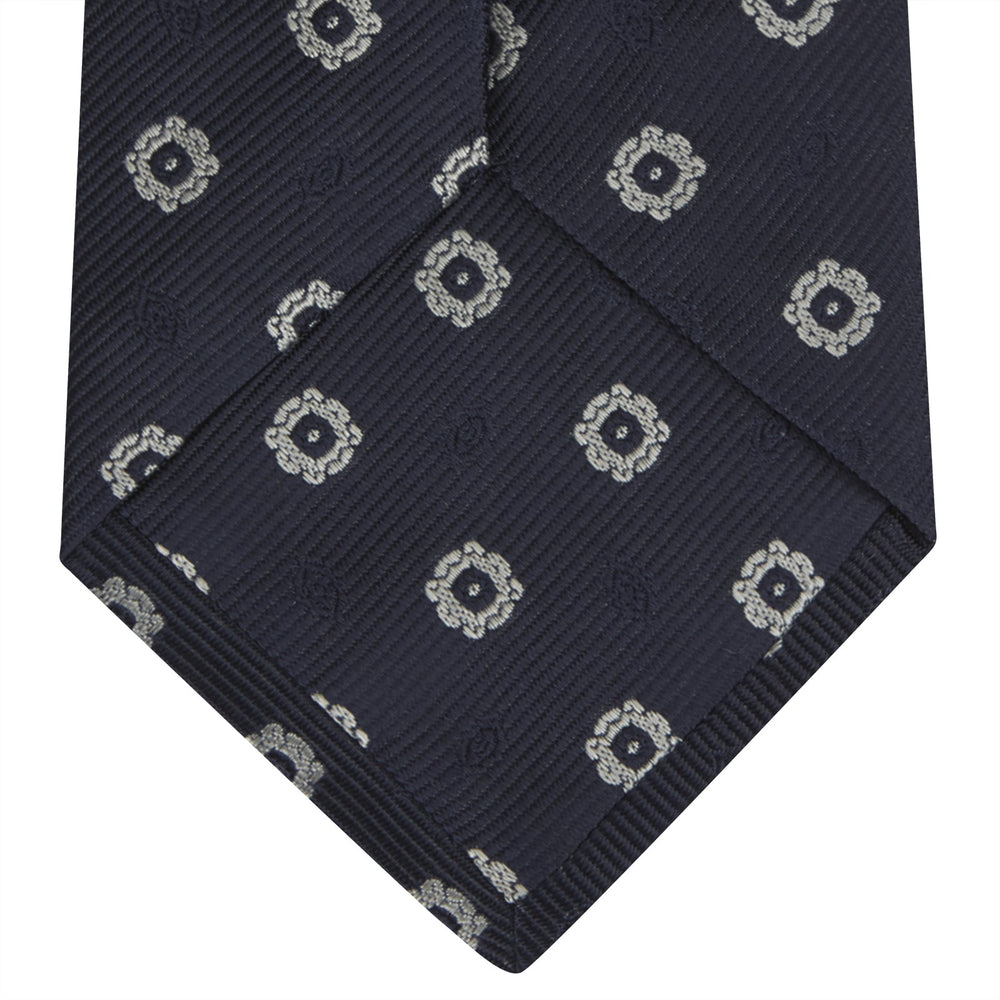 The Turnbull & Asser Navy and Silver Emblem Spot Silk Tie features a refined repeating emblem pattern in silver on navy silk, making it an elegant accessory for any office shirt.