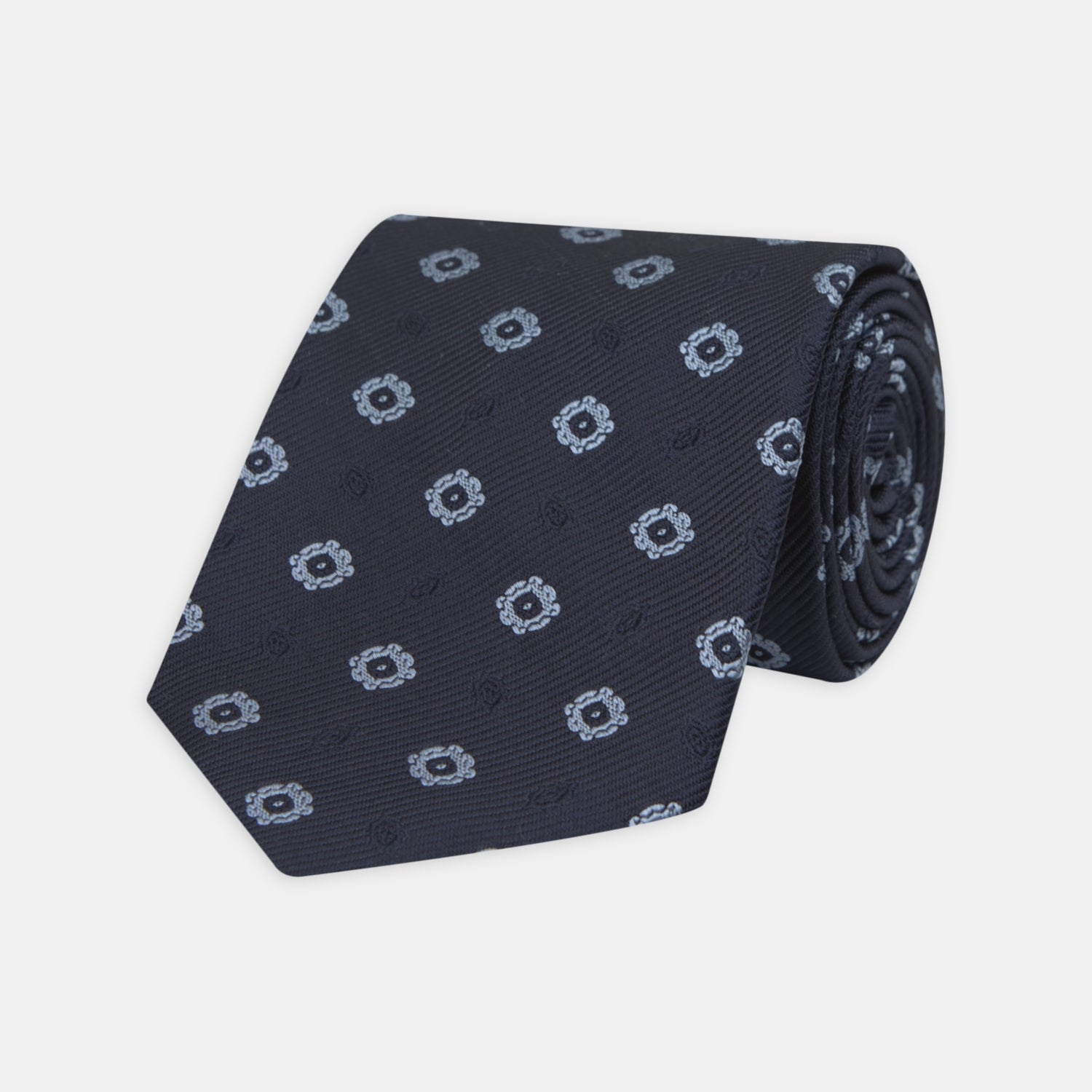 View 1 of product The Turnbull & Asser Navy and Light Blue Emblem Spot Silk Tie features a classic rolled design with a repeating light blue emblem pattern, ideal for office wear, shown against a plain white background.