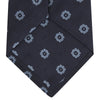 View 4 of product The Turnbull & Asser Navy and Light Blue Emblem Spot Silk Tie features a classic rolled design with a repeating light blue emblem pattern, ideal for office wear, shown against a plain white background.