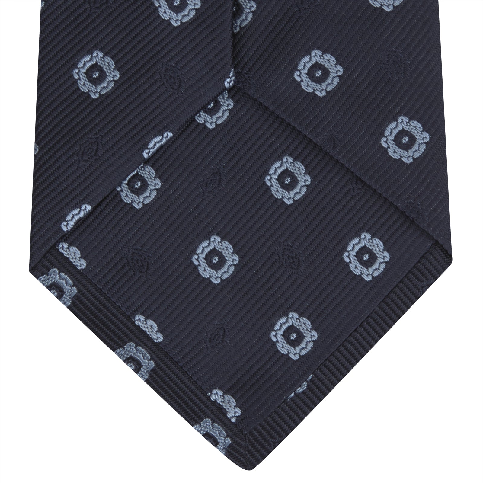 View 4 of product The Turnbull & Asser Navy and Light Blue Emblem Spot Silk Tie features a classic rolled design with a repeating light blue emblem pattern, ideal for office wear, shown against a plain white background.
