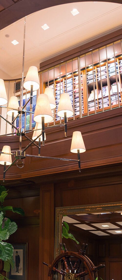 A large chandelier with multiple light shades hangs in a wood-paneled room. Above, shelves filled with neatly stacked folded shirts are visible behind a wooden railing.