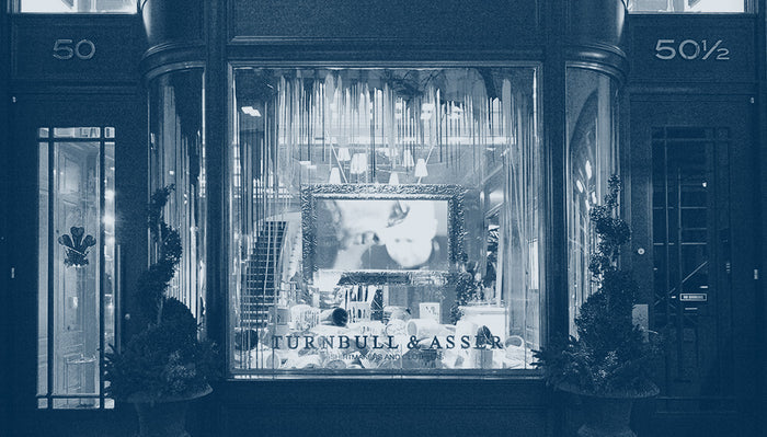 A shopfront at night displays a large window with Turnbull & Asser written on it. The interior is warmly lit, showing a framed picture and elegant decor. The address numbers 50 and 50½ are visible on either side of the entrance.