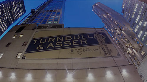 A low-angle view of tall buildings at dusk, with a large illuminated sign on one building reading Turnbull & Asser, Made in England next to an illustrated figure in a suit.