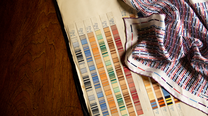 A fabric swatch book with rows of colored fabric samples lies open on a wooden surface next to a folded, patterned fabric featuring colorful, dashed stripes.
