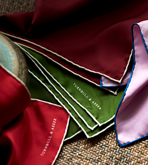 A close-up of colorful silk pocket squares in red, green, and lilac with white trim, all branded with Turnbull & Asser, displayed on a woven surface.