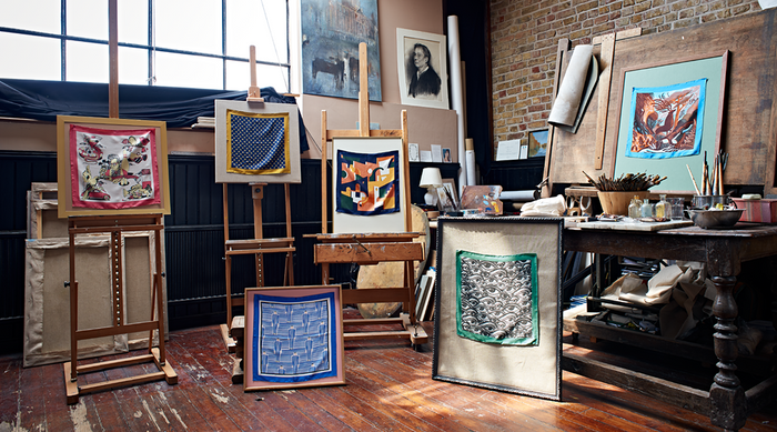 An artist’s studio with easels displaying colorful abstract art pieces, a wooden table holding brushes and art supplies, and natural light streaming through large windows onto a wooden floor.