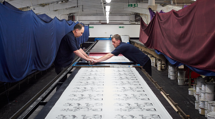 Two men work together in a textile factory, manually printing black-and-white designs onto a long sheet of fabric stretched on a large table, with colorful cloths draped along the sides.