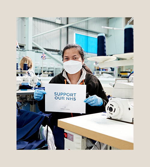 A woman wearing a mask and gloves sits in a textile factory, holding a sign that reads SUPPORT OUR NHS. Sewing machines and fabric are visible around her.