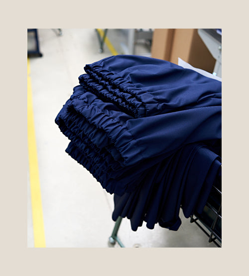 A stack of neatly folded navy blue garments with elastic waistbands rests on a metal cart in a brightly lit workspace with a yellow line on the floor nearby.