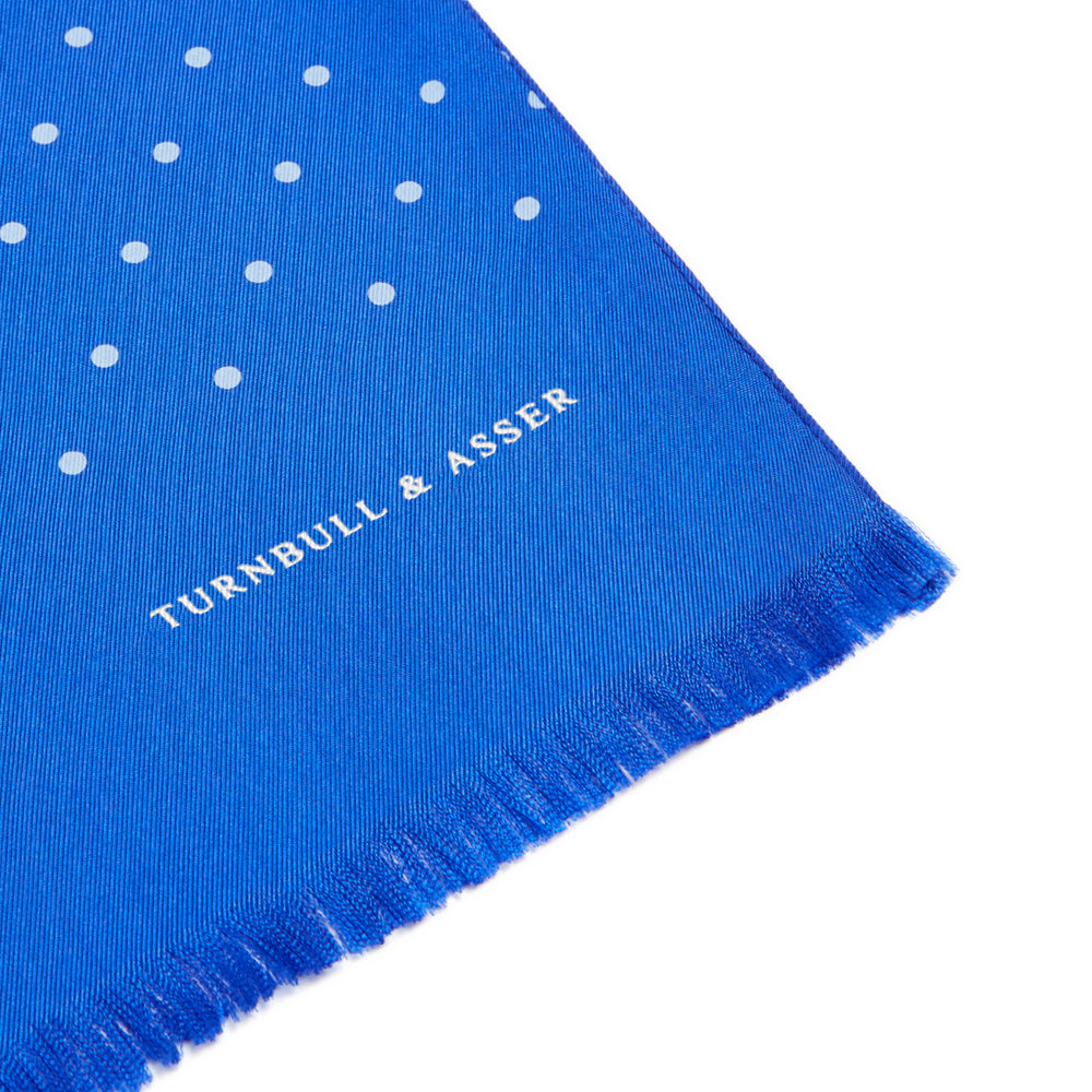 The Turnbull & Asser Royal Blue and White Spotted Silk Scarf features a soft silk fabric, short fringes at both ends, neatly folded center, and a hand-printed polka dot design for a distinctive, elegant look.