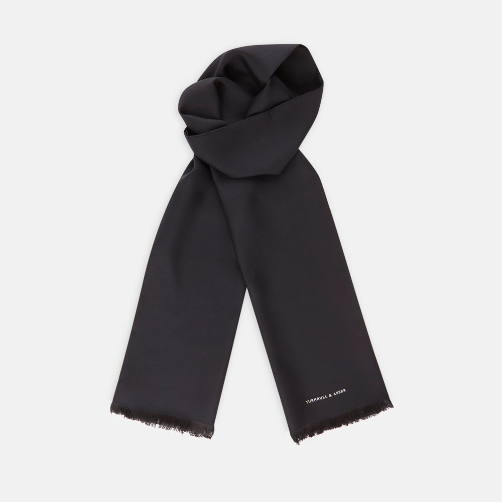 The Turnbull & Asser Plain Black Silk Scarf features fringed edges and subtle white branding at one end, ideal for formalwear or tuxedos, and is shown on a plain white background.