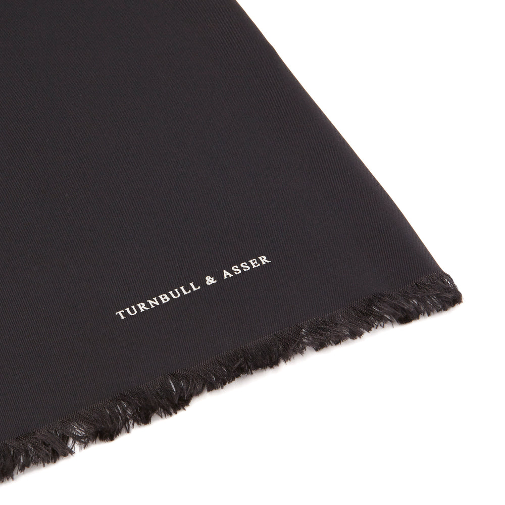 The Turnbull & Asser Plain Black Silk Scarf features fringed edges and subtle white branding at one end, ideal for formalwear or tuxedos, and is shown on a plain white background.