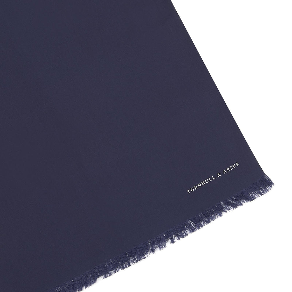 A Plain Navy Silk Scarf by Turnbull & Asser, neatly folded with both ends hanging down, is shown against a white background. One end displays subtle white branding, highlighting this elegant accessory.