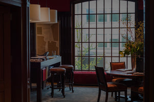 A cozy, dimly lit room with a round table and chairs near a large window. Bar stools line a counter under hanging lights, and leafy branches sit in a vase by the window with city buildings visible outside.