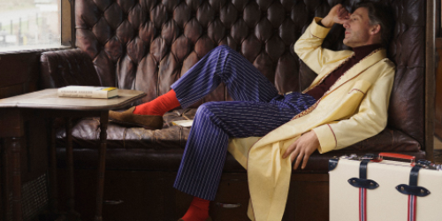 A man in a yellow robe, red socks, and blue striped pants relaxes on a tufted leather bench inside a train car, with a book on the table and a white suitcase nearby.