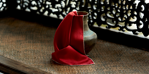A bronze vase holds a folded red silk handkerchief, set on a woven surface in front of an ornate, dark wooden screen with intricate patterns.