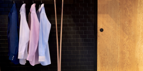 Four dress shirts in blue, white, pink, and light blue are hanging on a rack against a black tiled wall next to a wooden door.