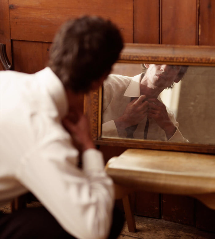 A person in a white shirt adjusts their collar while looking into an old, rectangular mirror on a wooden table, with their reflection clearly visible.