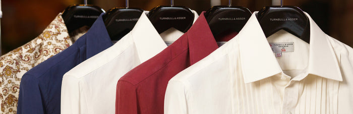 Five neatly hung dress shirts on black hangers, including white, red, blue, and a patterned shirt, displayed in a row. The hangers are labeled Turnbull & Asser.