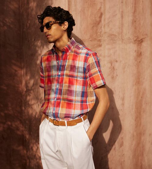 A person wearing sunglasses, a colorful plaid short-sleeve shirt, and white pants with a tan belt stands against a textured brown wall, looking to the side.