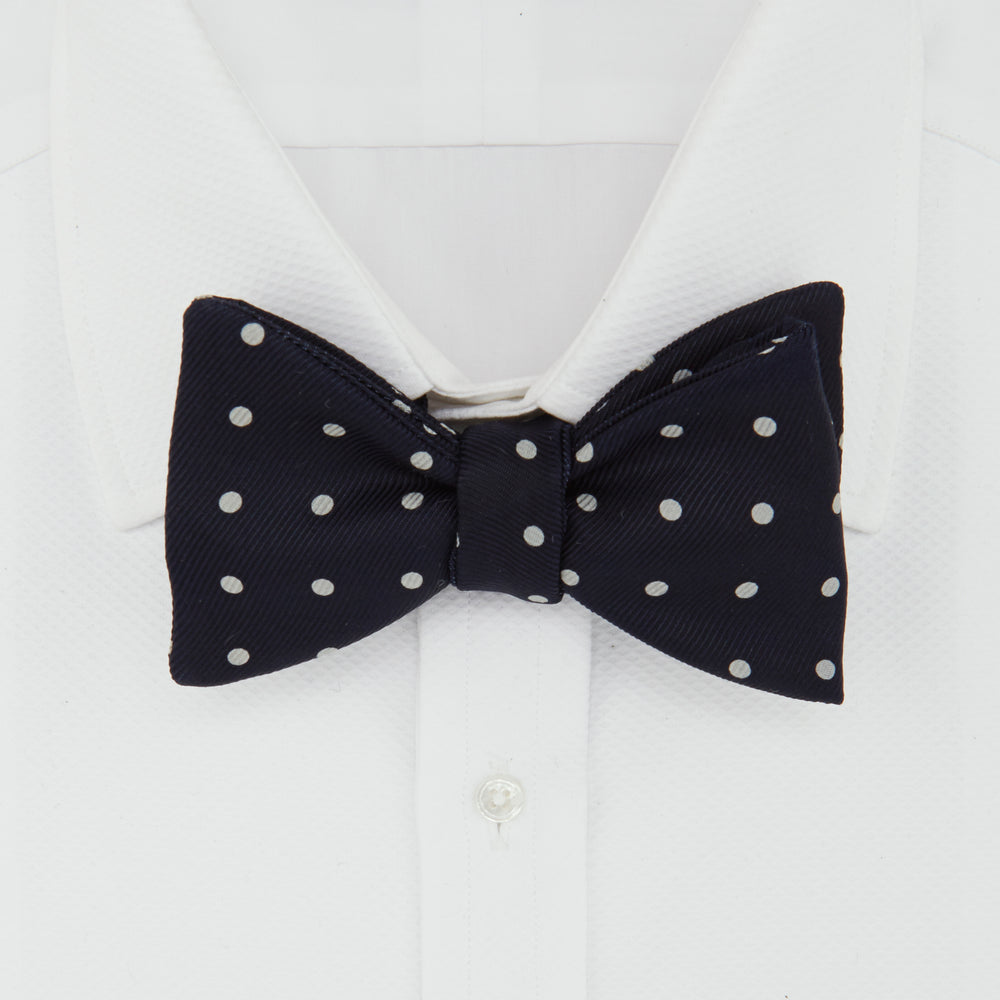 The Winston Navy and White Spot Silk Bow Tie by Turnbull & Asser features classic white polka dots on navy silk, reminiscent of Winston Churchill's style, and is shown against a plain white background.
