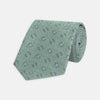 View 1 of product The Turnbull & Asser Green Star Flowers Silk Tie features a light green jacquard with a subtle checkered pattern and small blue star flower motifs, shown rolled up on a white background.