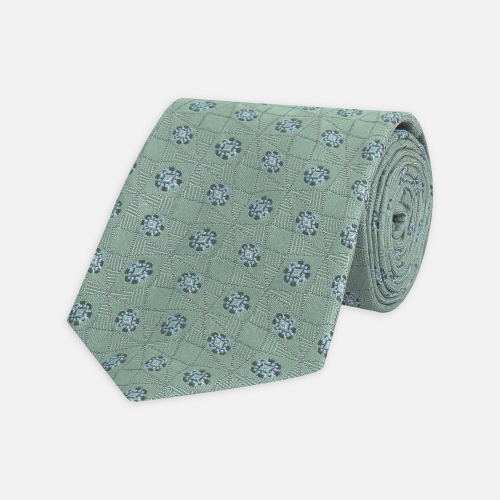 The Turnbull & Asser Green Star Flowers Silk Tie features a light green jacquard with a subtle checkered pattern and small blue star flower motifs, shown rolled up on a white background.