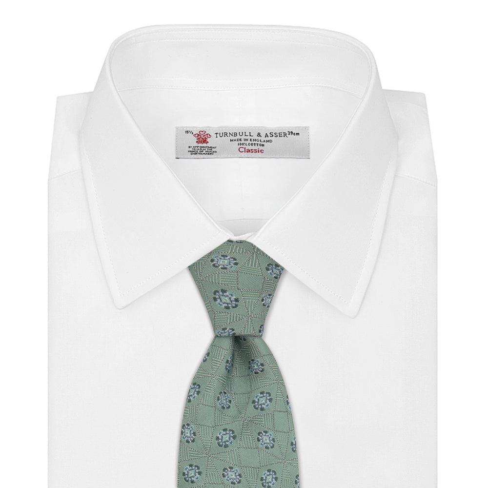 The Turnbull & Asser Green Star Flowers Silk Tie features a light green jacquard with a subtle checkered pattern and small blue star flower motifs, shown rolled up on a white background.