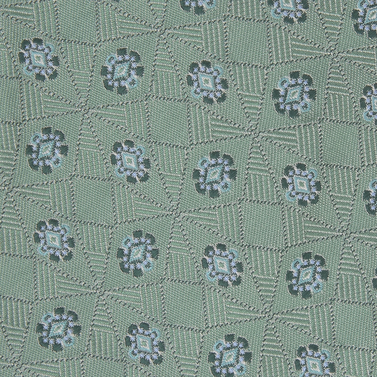 View 3 of product The Turnbull & Asser Green Star Flowers Silk Tie features a light green jacquard with a subtle checkered pattern and small blue star flower motifs, shown rolled up on a white background.