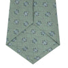 View 4 of product The Turnbull & Asser Green Star Flowers Silk Tie features a light green jacquard with a subtle checkered pattern and small blue star flower motifs, shown rolled up on a white background.