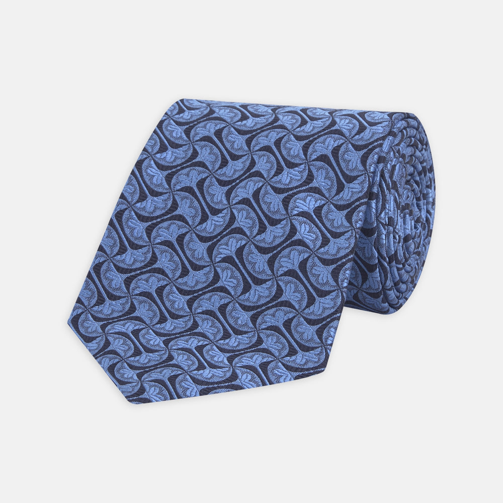 The Turnbull & Asser Blue Geo Silk Tie features a refined blue silk with a light blue abstract leaf pattern in a geometric design, rolled at one end and shown on a plain white background.