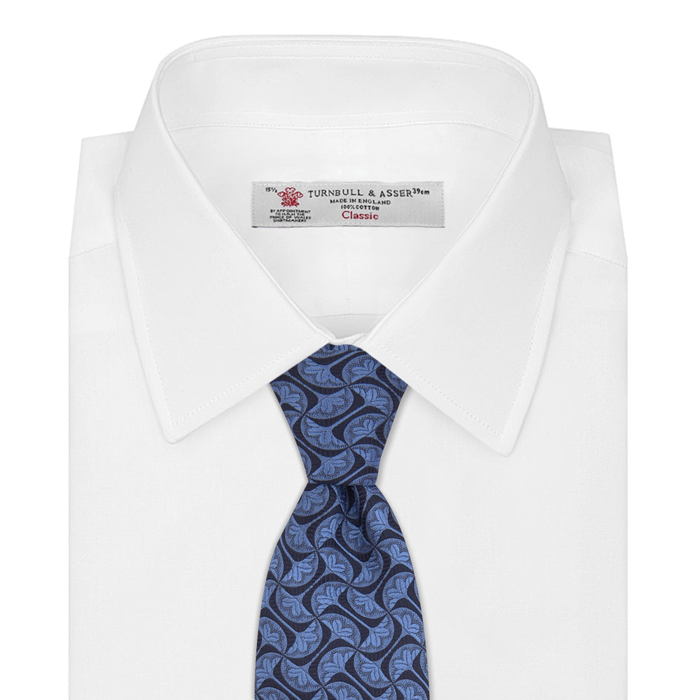 The Turnbull & Asser Blue Geo Silk Tie features a refined blue silk with a light blue abstract leaf pattern in a geometric design, rolled at one end and shown on a plain white background.