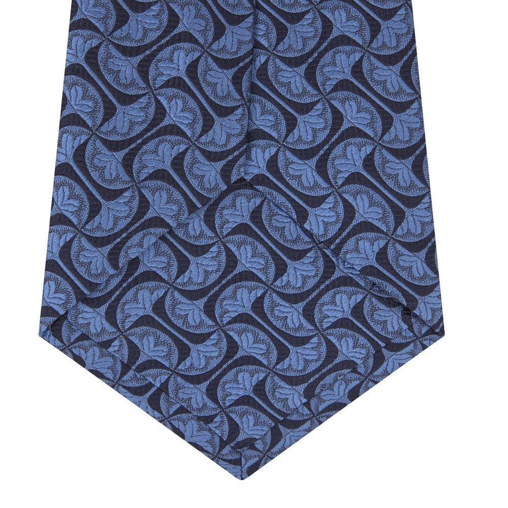 The Turnbull & Asser Blue Geo Silk Tie features a refined blue silk with a light blue abstract leaf pattern in a geometric design, rolled at one end and shown on a plain white background.