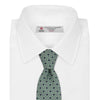 View 2 of product The Leaf Green Geometric Silk Tie by Turnbull & Asser features a repeating pattern of small navy blue and white floral shapes, evoking a mid-century motif. It is neatly rolled and displayed on a white background.