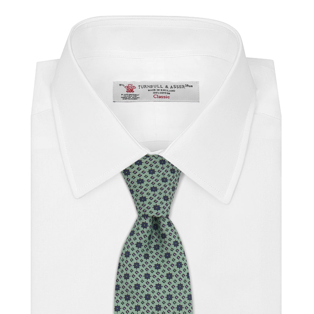 The Leaf Green Geometric Silk Tie by Turnbull & Asser features a repeating pattern of small navy blue and white floral shapes, evoking a mid-century motif. It is neatly rolled and displayed on a white background.