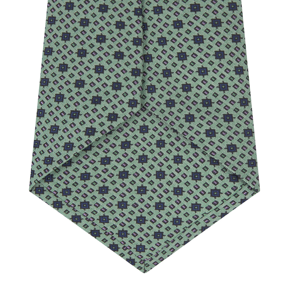The Leaf Green Geometric Silk Tie by Turnbull & Asser features a repeating pattern of small navy blue and white floral shapes, evoking a mid-century motif. It is neatly rolled and displayed on a white background.
