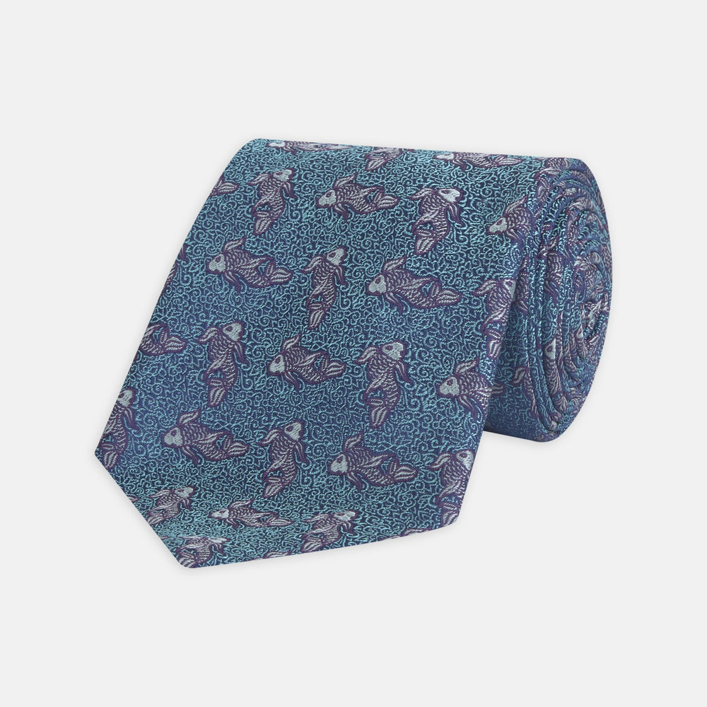 The Turnbull & Asser Turquoise Carp Silk Tie is woven in England, featuring a repeating white and brown carp design on a light blue background with intricate swirling patterns.