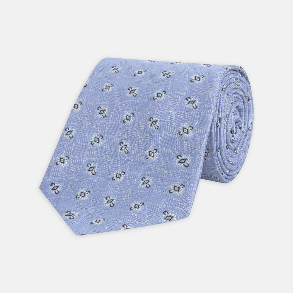 The Turnbull & Asser Sky Blue Star Flowers Silk Tie features a small white and dark blue square and star flower pattern on jacquard silk, shown rolled up against a plain white background.
