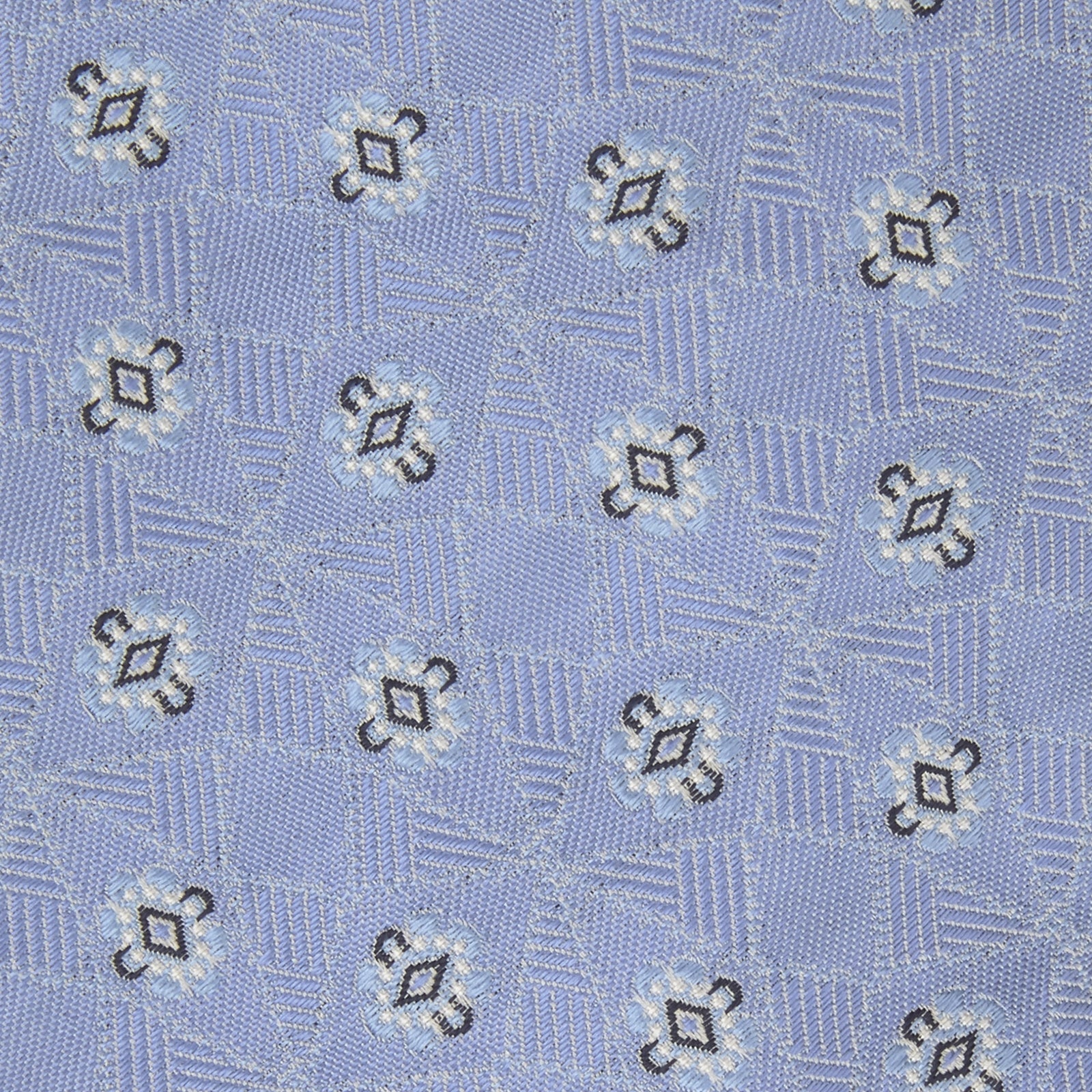 View 3 of product The Turnbull & Asser Sky Blue Star Flowers Silk Tie features a small white and dark blue square and star flower pattern on jacquard silk, shown rolled up against a plain white background.