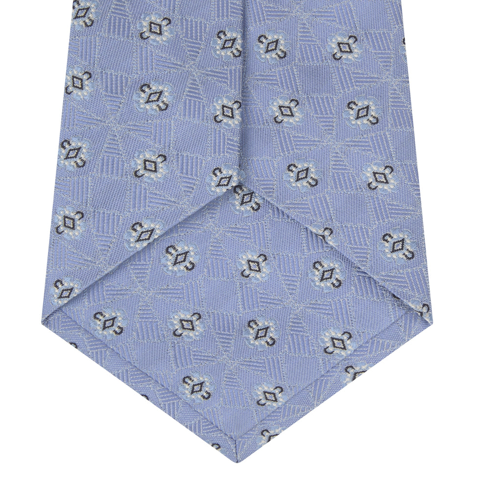 The Turnbull & Asser Sky Blue Star Flowers Silk Tie features a small white and dark blue square and star flower pattern on jacquard silk, shown rolled up against a plain white background.