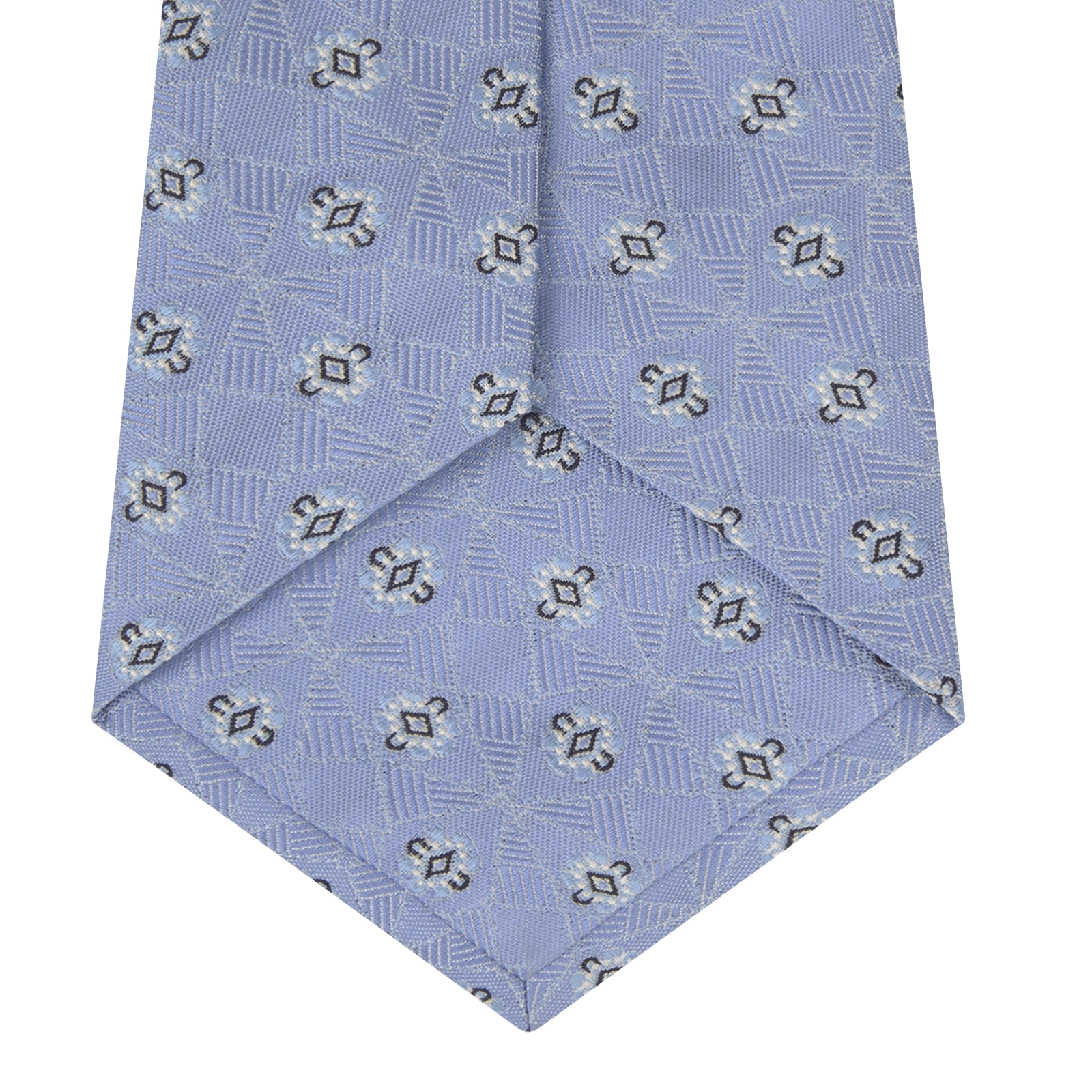 View 4 of product The Turnbull & Asser Sky Blue Star Flowers Silk Tie features a small white and dark blue square and star flower pattern on jacquard silk, shown rolled up against a plain white background.