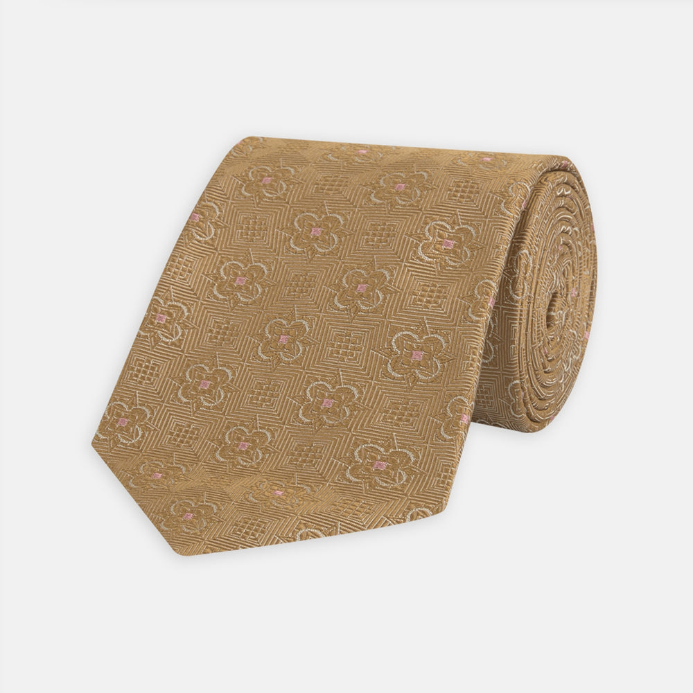 The Gold Flower Mosaic Silk Tie by Turnbull & Asser features an intricate gold jacquard pattern of geometric shapes and floral designs in pink and light brown hues.