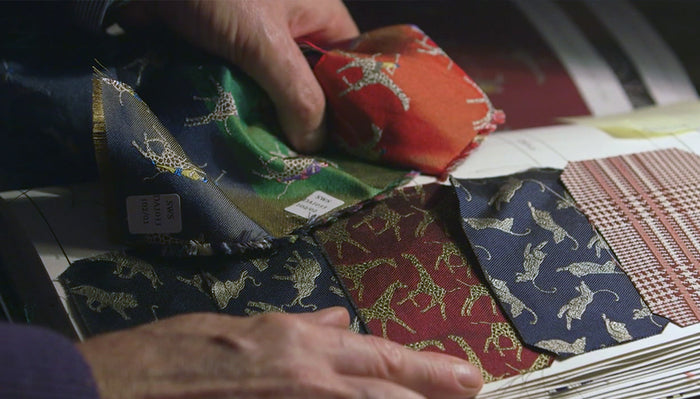 Close-up of hands holding and examining colorful fabric swatches with animal patterns, laid out on an open book or sample binder. The fabrics feature designs of leaping animals in various colors.