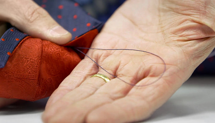 A close-up of a hand holding a threaded needle, with a red patterned fabric and a blue fabric with red dots. The hand is wearing a gold ring.